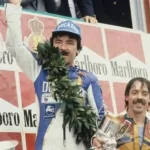 Ángel Nieto 12+1 celebrando una victoria en el podio con un casco de carreras y una corona de laurel, rodeado de patrocinadores.