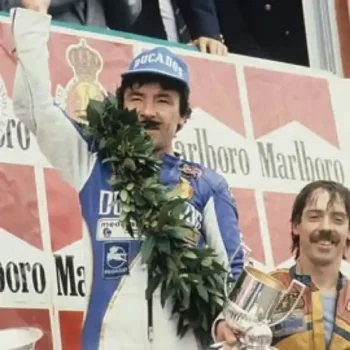 Ángel Nieto 12+1 celebrando una victoria en el podio con un casco de carreras y una corona de laurel, rodeado de patrocinadores.