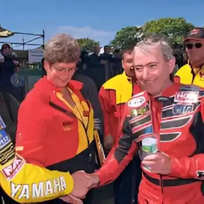 Joey Dunlop Legado Joey Dunlop, con una sonrisa, estrechando la mano de otra persona mientras sostiene una botella, simbolizando su legado y conexión con la gente.